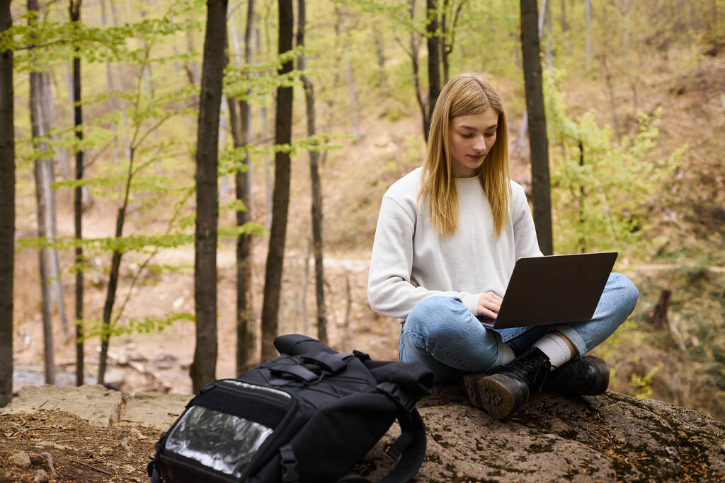 Молодая блондинка с рюкзаком прогулки в лесу, сидя на валуне с ноутбуком и работы - Фото, изображение