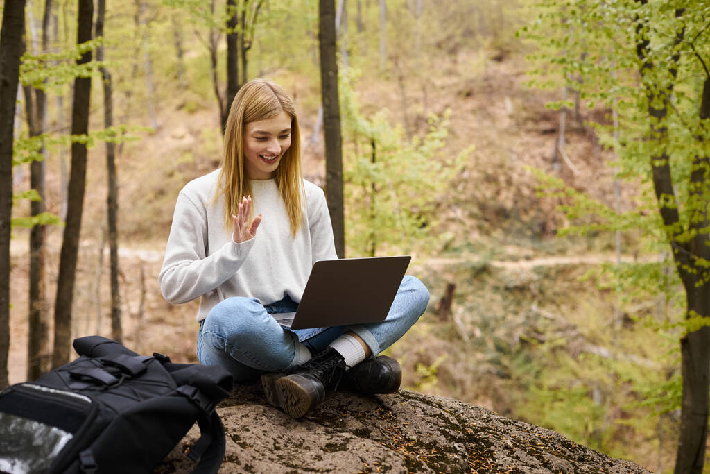 Молодая блондинка с рюкзаком в лесу, сидит на валуне с ноутбуком и делает видеозвонки - Фото, изображение