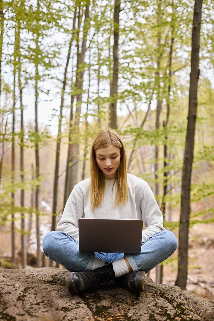 blond relaxed woman on halt in woods, sitting on boulder with laptop in lotus position - Photo, Image