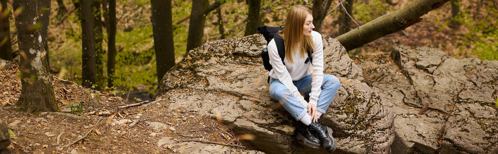 Mladá žena turistka s batohem sedí na místě na skalnatém útesu v lese dívá pryč, prapor - Fotografie, Obrázek