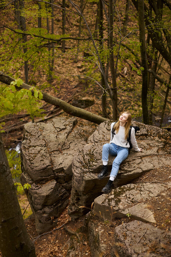 Giovane escursionista donna con zaino seduto a fermarsi sulla scogliera rocciosa nella foresta guardando nel cielo - Foto, immagini