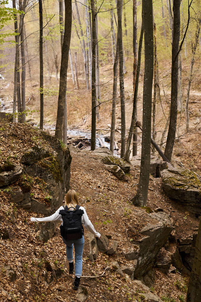 Back view of young blonde female traveler wearing backpack running to forest stream - Photo, Image