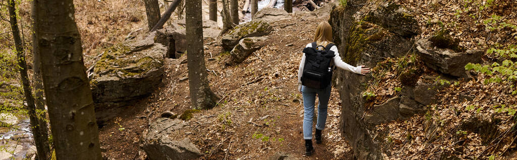 Back view of young blonde female traveler wearing backpack holding trees near forest river, banner - Photo, Image