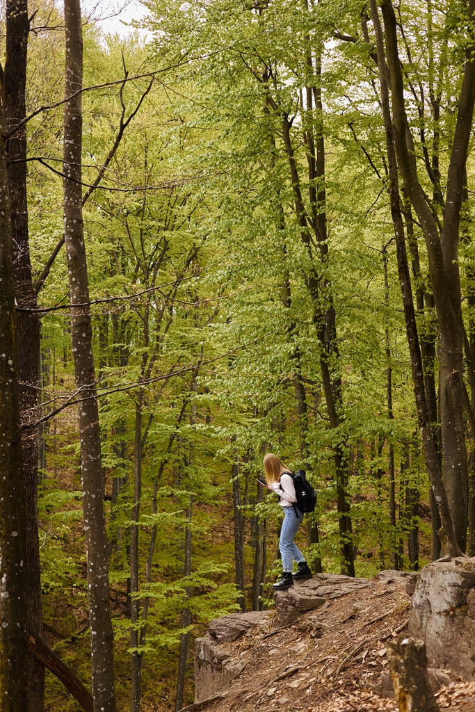 Vista lateral da mulher loira curiosa usando mochila em caminhadas e caminhadas em penhasco rochoso com telefone - Foto, Imagem