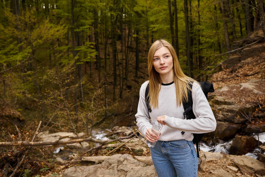 Pretty blonde female hiker wearing sweater and jeans crossing the forest creek walking in woods - Photo, Image