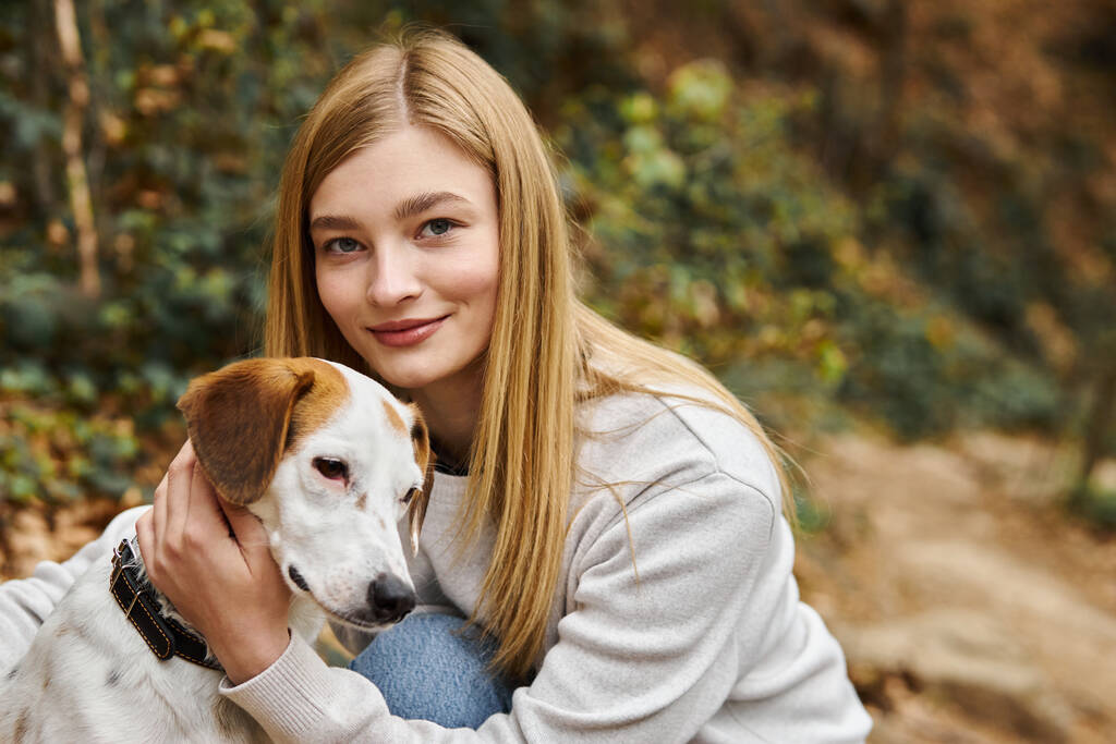 Blonde Frau liebt und umarmt sanft ihren Hund und schaut beim Gassigehen im Wald in die Kamera - Foto, Bild