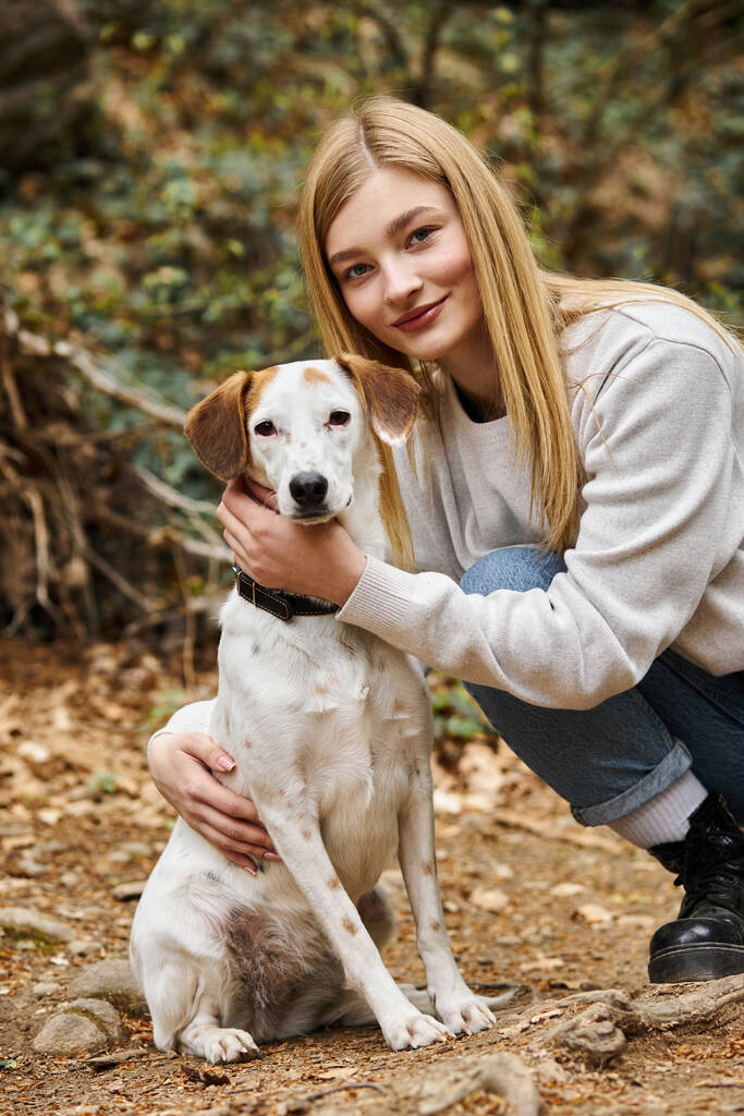Cheerful woman loving and hugging gently her dog and looking at camera while walking in woods - Photo, Image