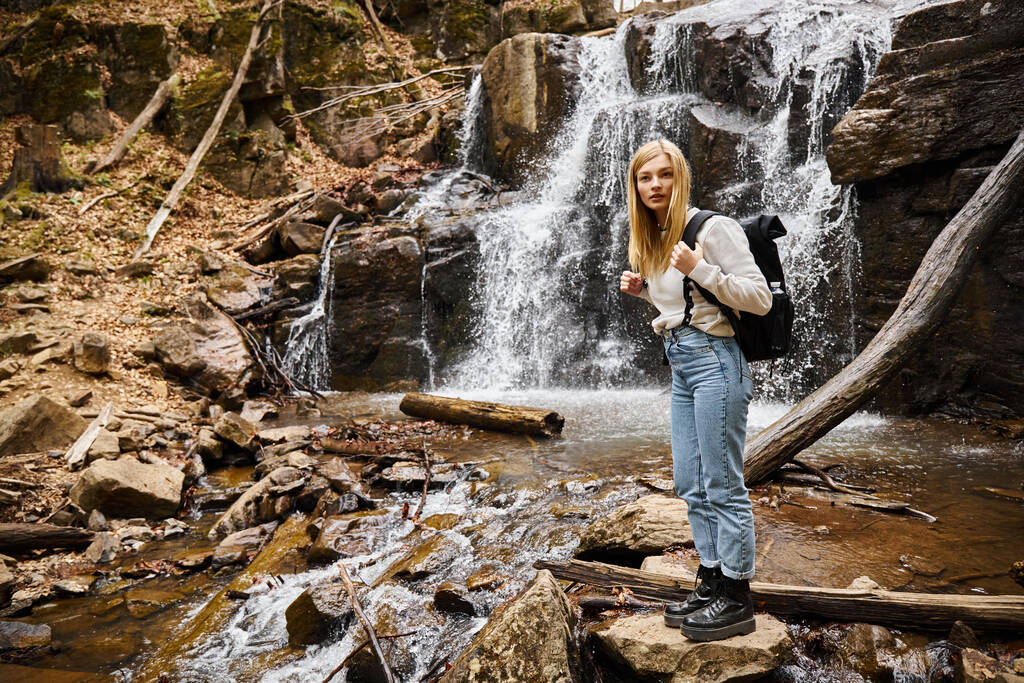Active blonde female hiker standing by the forest creek walking on rocks near waterfall - Photo, Image