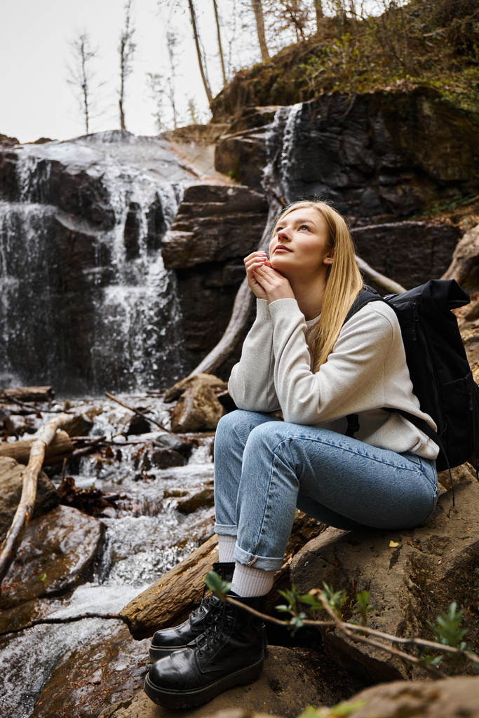 Dreamy woman sitting with backpack and looking in the sky in forest near waterfall - Photo, Image