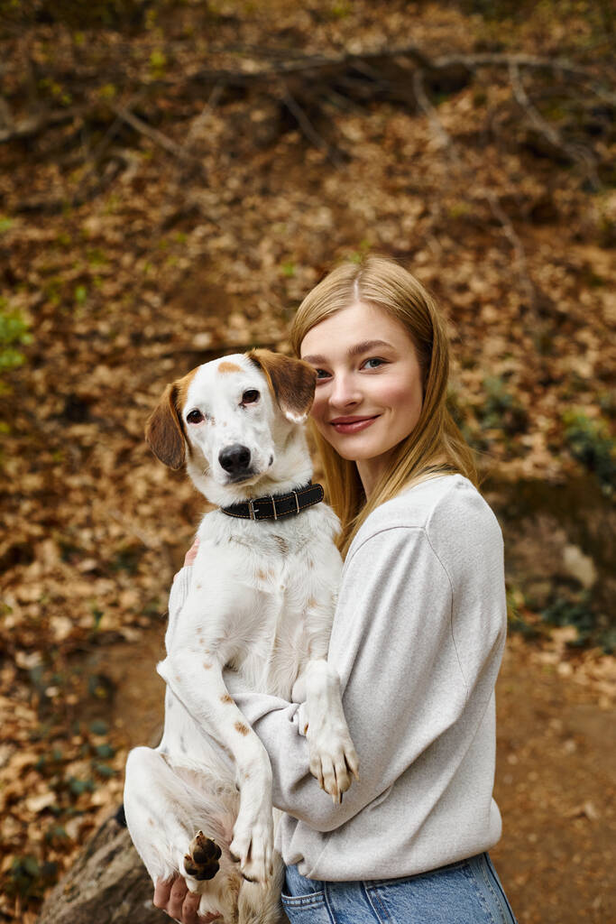 Mujer joven sosteniendo a su perro en las manos y abrazándose en el descanso de senderismo con vista a la montaña y el bosque - Foto, imagen