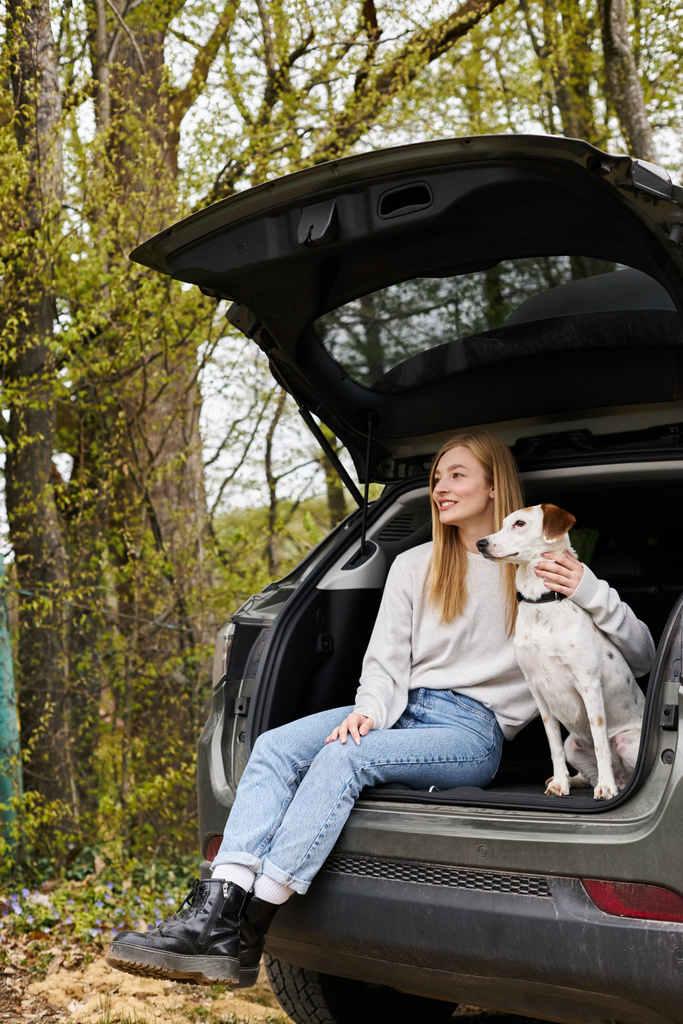 Nyugodt nő ölelgeti a kutyáját a kocsi hátuljában ülve, és félrenéz az erdőben túrázni. - Fotó, kép