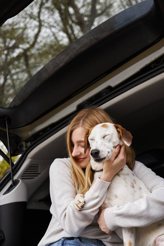 Улыбающаяся счастливая женщина, обнимающая собаку, сидящую на заднем сиденье машины в лесу во время похода, останавливается, глаза закрываются - Фото, изображение