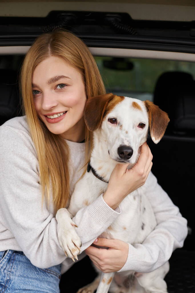 笑顔の幸せな女性は,森の中で車の後ろに座ってカメラを見ている彼女の犬を抱きしめます - 写真・画像