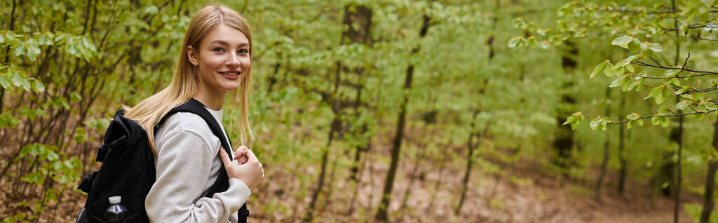 Powrót widok portret uśmiechniętej blondynki podróżującej po lesie i noszącej plecak, baner - Zdjęcie, obraz
