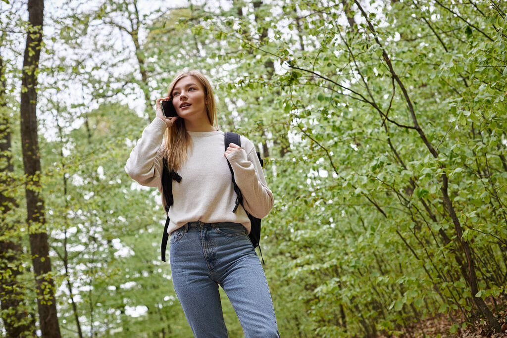 Photo of blond pretty woman traveler with backpack talking by phone walking in forest path - Photo, Image