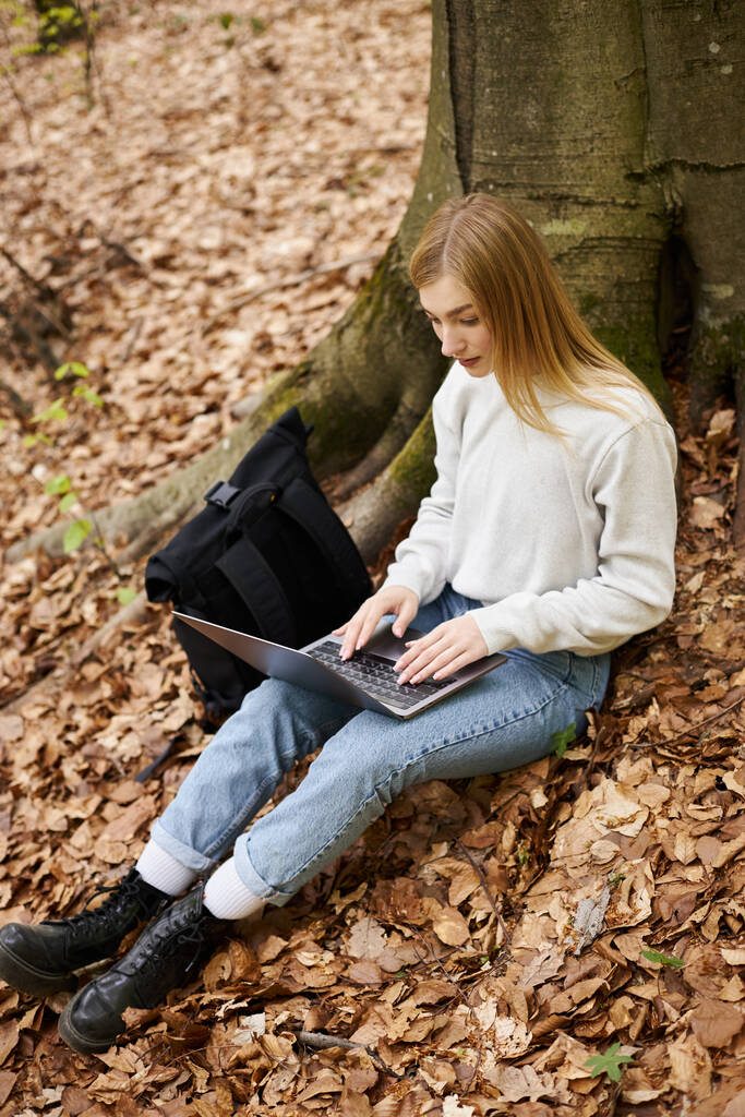 Relaxed blonde woman with laptop on her legs working remote while sitting in forest on trip - Photo, Image