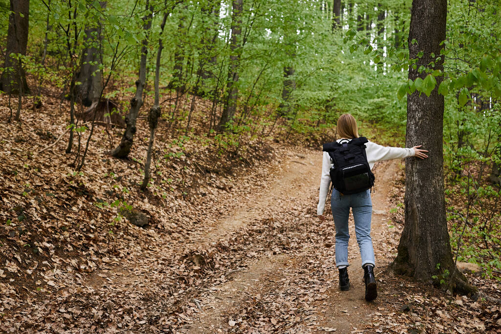 Back full length view of blonde woman hiking walking in green forest touching trees - Photo, Image