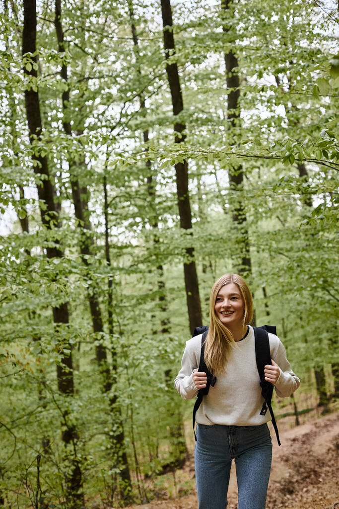 Smiling hiking blonde woman wearing sweater and backpack walking in forest scenery in woods - Photo, Image