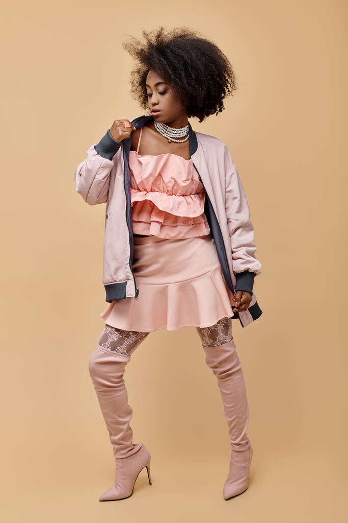 menina americana africana na década de 20, posando em roupa de pêssego com jaqueta e botas sobre o joelho no bege, 2024 - Foto, Imagem