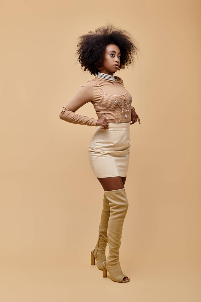 Chica afroamericana de moda en mini falda pastel y botas altas en un telón de fondo beige - Foto, imagen
