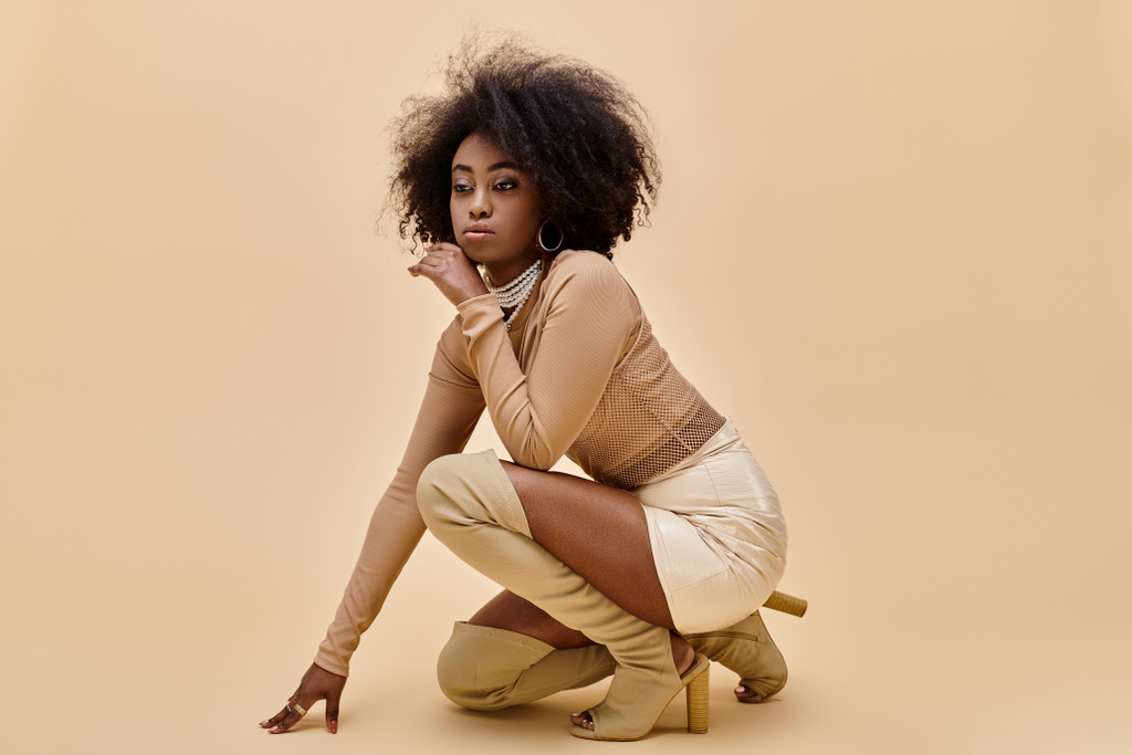 dromerig Afrikaans Amerikaans model in stijlvolle outfit en dijhoge laarzen poseren op beige achtergrond - Foto, afbeelding