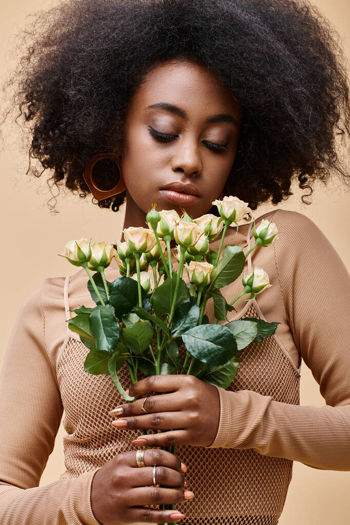 joven y rizada mujer afroamericana mirando pequeñas rosas sobre fondo beige, color pelusa melocotón - Foto, imagen