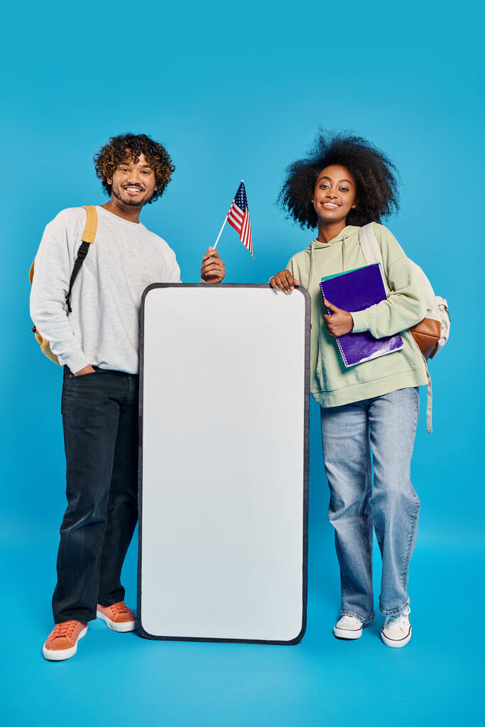 Ένα ζευγάρι διαφορετικών ατόμων, πολυπολιτισμικοί φοιτητές, στέκονται κοντά σε ένα mockup smartphone σε ένα στούντιο σε ένα μπλε φόντο. - Φωτογραφία, εικόνα