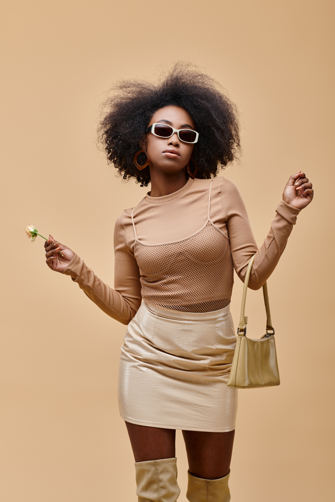 κομψό αφρικανικό αμερικανικό μοντέλο σε γυαλιά ηλίου κρατώντας τσάντα και μικροσκοπικό τριαντάφυλλο σε μπεζ φόντο - Φωτογραφία, εικόνα