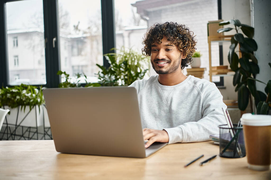 Um homem de fundo diverso senta-se atentamente na frente de um computador portátil em um espaço de co-trabalho moderno, engajando-se em trabalho digital. - Foto, Imagem