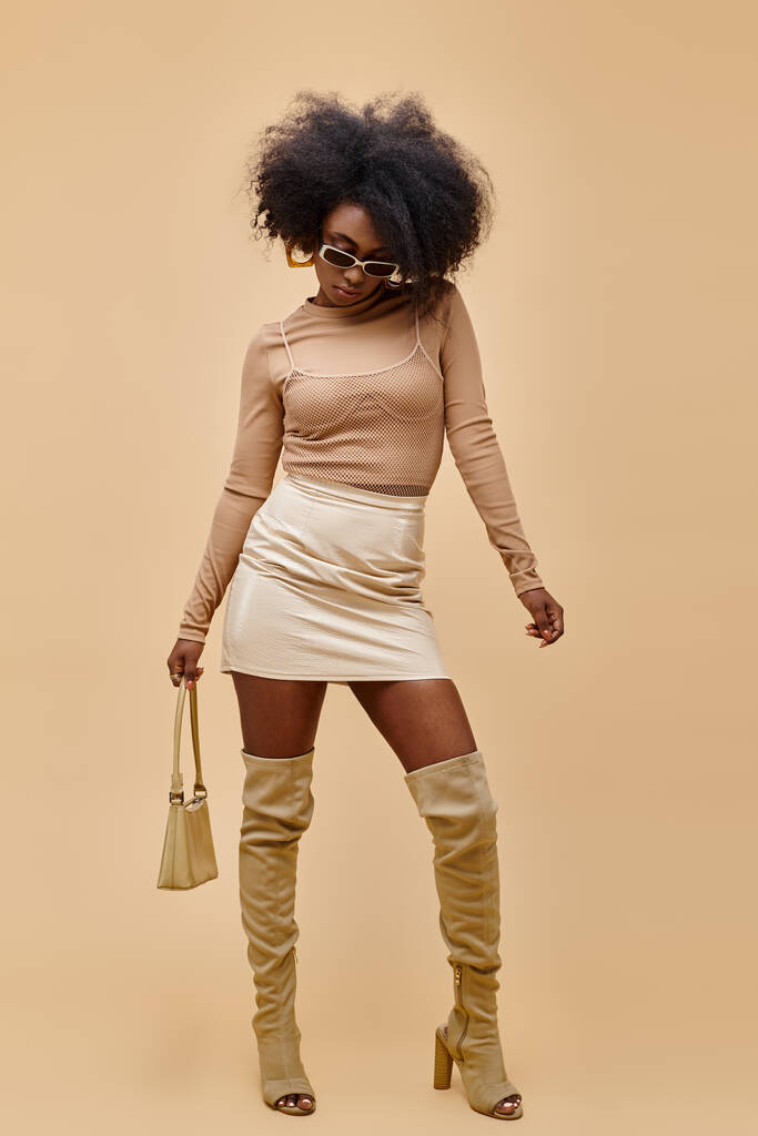 modelo afroamericano en gafas de sol y botas de muslo alto sosteniendo bolso de moda sobre fondo beige - Foto, imagen