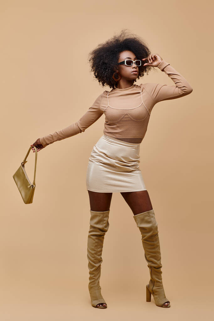 donna afroamericana in occhiali da sole e stivali alti che tengono la borsa su sfondo beige - Foto, immagini