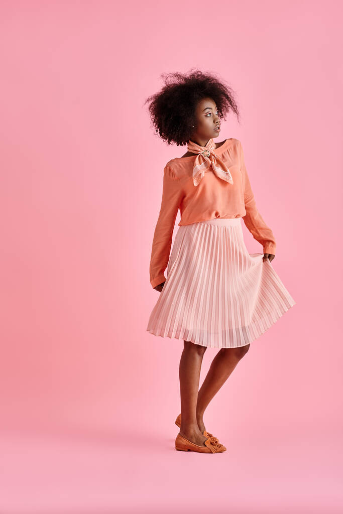 молода афроамериканка в персиковій блузці і спідниці міді вдарила позу на пастельному рожевому фоні - Фото, зображення
