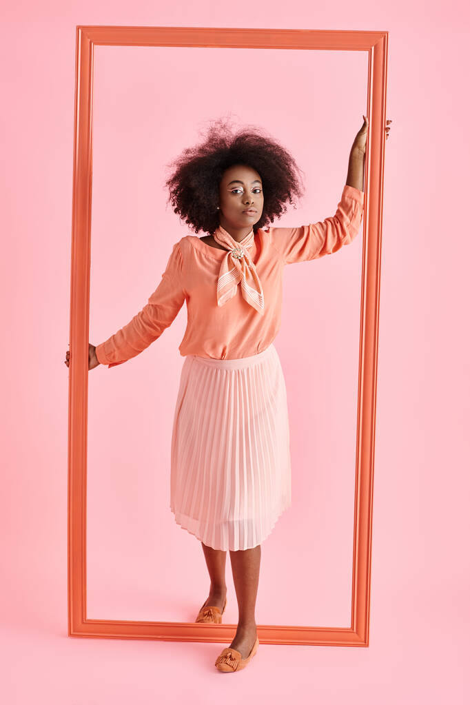 pretty african american woman in peach blouse and midi skirt strikes pose near frame on pastel pink - Photo, Image