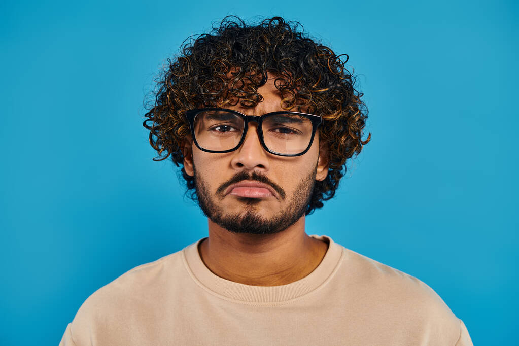 Индийский студент в кудрявых волосах и очках уверенно позирует на синем фоне в студии. - Фото, изображение