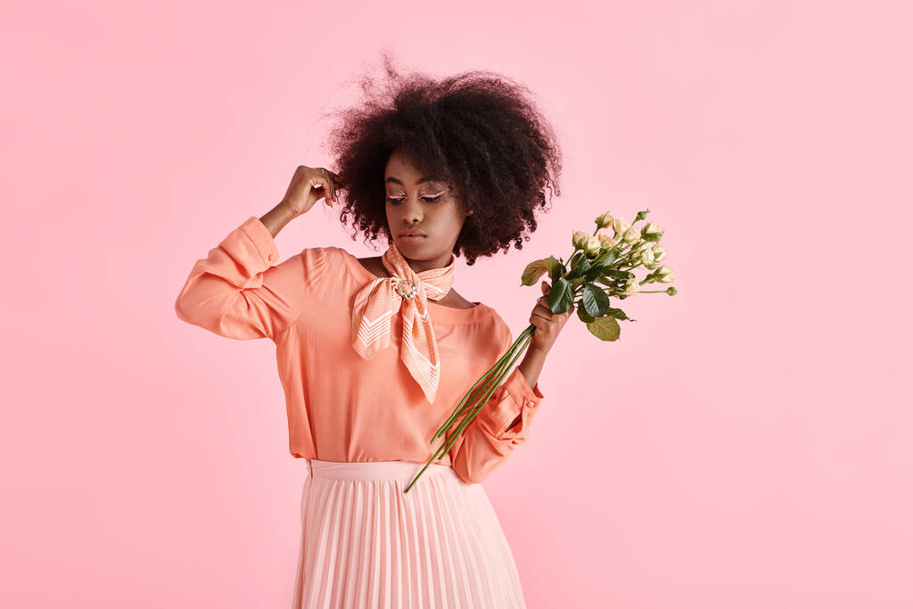 афроамериканська жінка в одязі з персиковими фузами, що тримає квіти і дивиться вниз на рожевому фоні - Фото, зображення