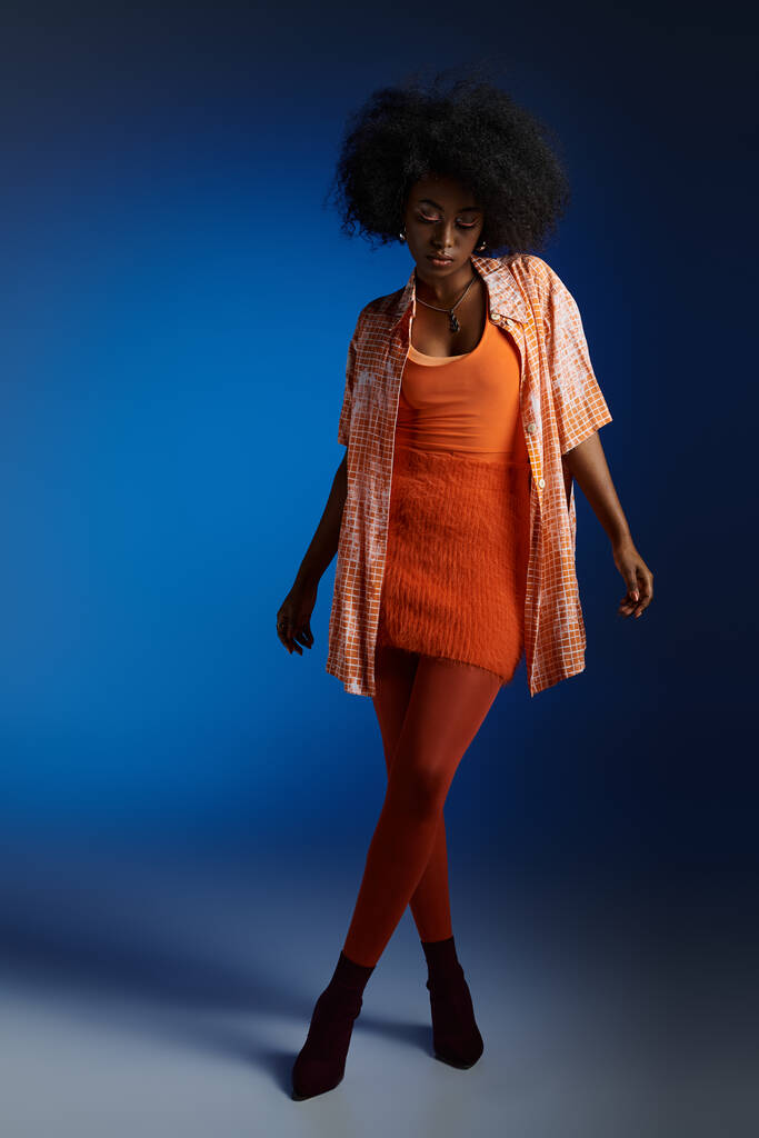 Шикарний вигляд молодої афроамериканки у візерунковій сорочці та помаранчевій сукні на синьому фоні - Фото, зображення