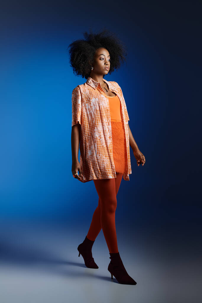 Aparência chique de menina americana africana bonita em camisa estampada e vestido laranja no fundo azul - Foto, Imagem