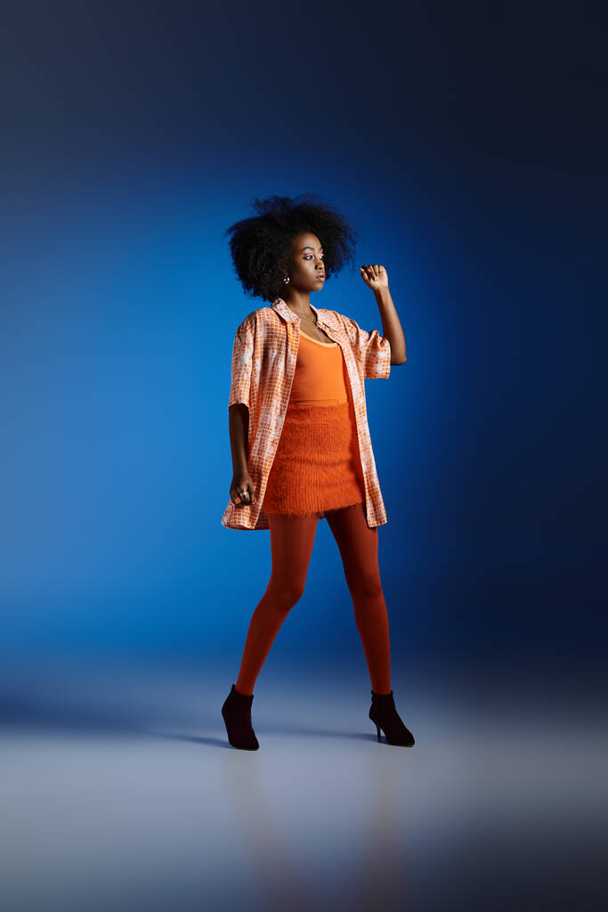 elegante look de mujer afroamericana en camisa estampada y vestido naranja posando sobre fondo azul - Foto, imagen