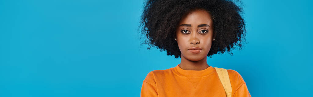 Stylowa Afroamerykanka w luźnym stroju stoi pewnie z jej obszernymi afro włosami, przed tętniącym życiem niebieskim tłem. - Zdjęcie, obraz