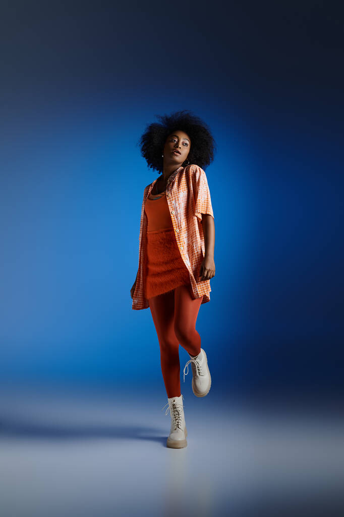 módní vzhled afrického amerického modelu v vzorované košili a oranžové šaty pózují na modrém pozadí - Fotografie, Obrázek