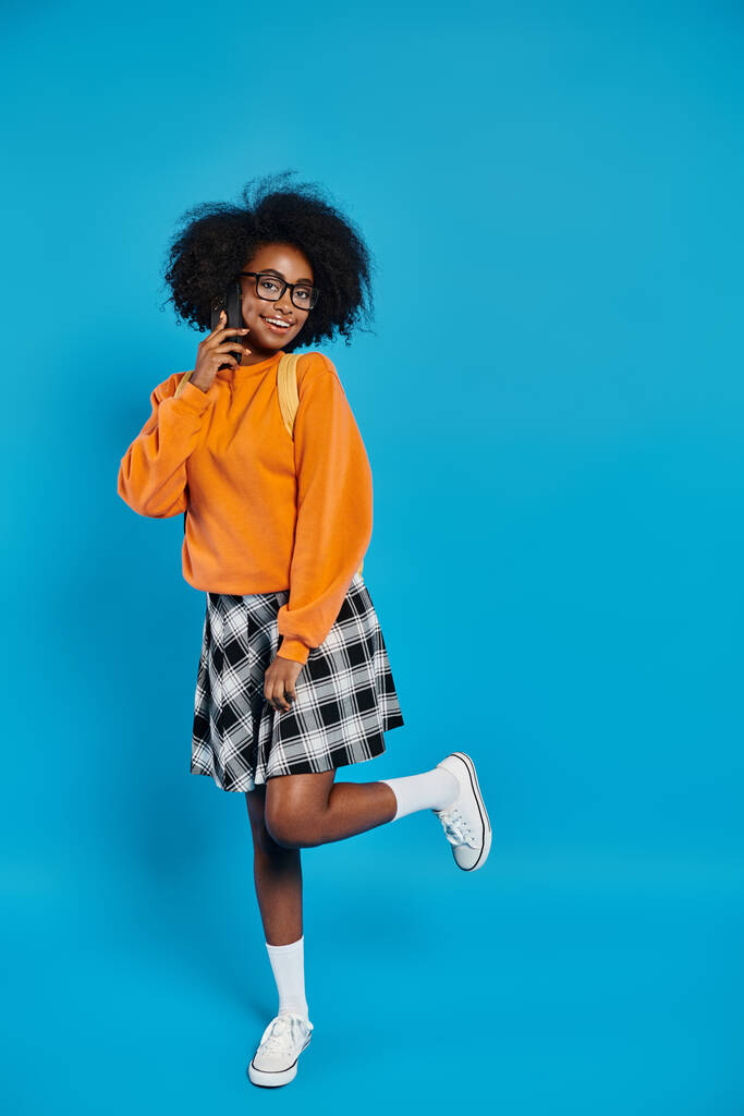 Une étudiante afro-américaine élégante se tient avec confiance dans un pull orange vif et une jupe à carreaux tendance sur fond bleu. - Photo, image