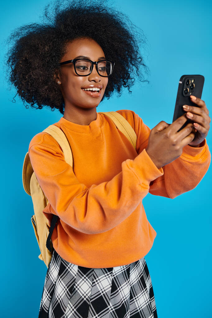 Μια νεαρή Αφρο-Αμερικανίδα με απλή ενδυμασία βγάζει μια σέλφι με το κινητό της ενώ κουβαλάει ένα σακίδιο, σε ένα μπλε φόντο σε ένα στούντιο.. - Φωτογραφία, εικόνα