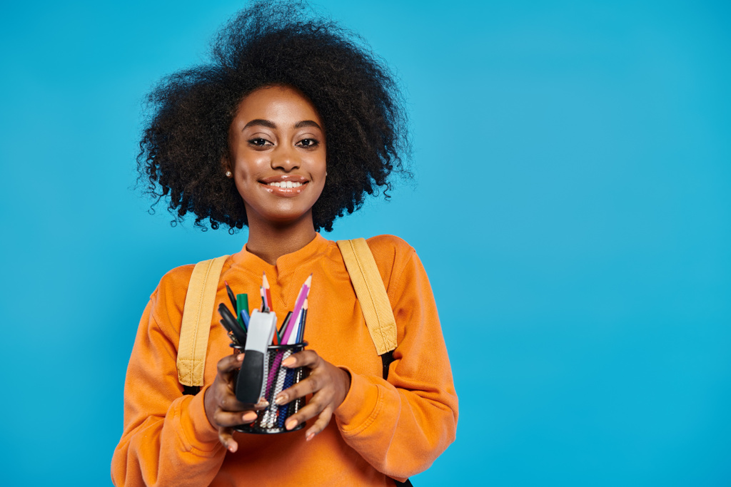 Une étudiante afro-américaine en tenue décontractée tient une tasse remplie d'un assortiment de stylos et crayons sur fond bleu. - Photo, image