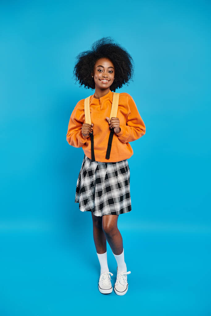 Uma menina universitária afro-americana fica confiante em trajes casuais contra um cenário azul vibrante em um cenário de estúdio. - Foto, Imagem