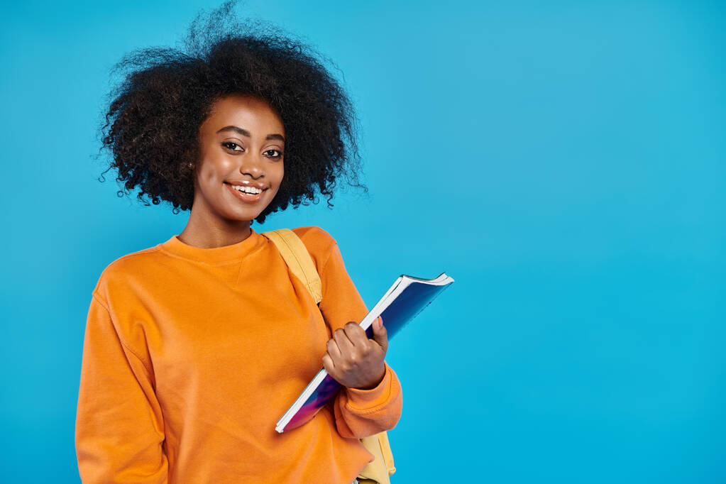 Una joven universitaria afroamericana de pie con atuendo casual, sosteniendo un libro, sonriendo calurosamente a la cámara con un telón de fondo azul. - Foto, Imagen