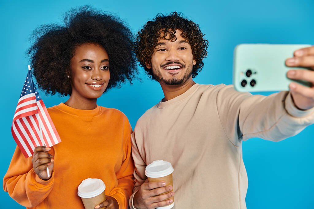 An interracial man and woman in casual attire take a selfie with American flag against a blue backdrop in a studio. - Photo, Image