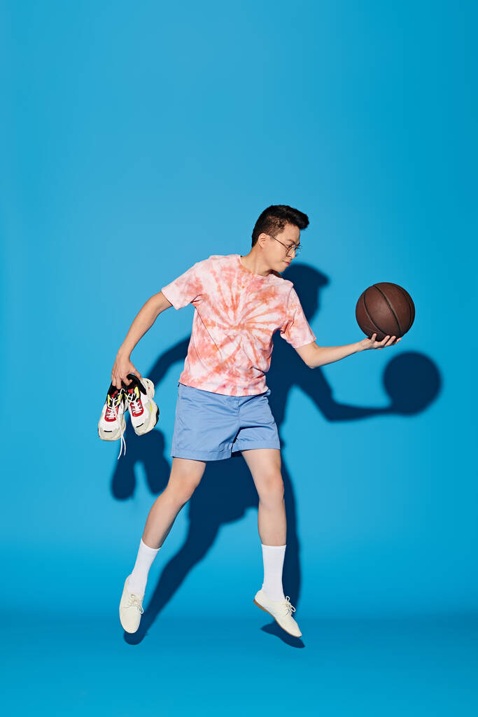 Modayı takip eden genç bir adam canlı mavi bir arka planda basketbol topuyla poz veriyor.. - Fotoğraf, Görsel