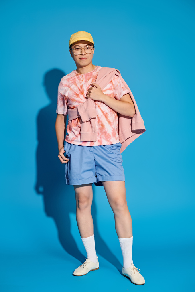 Mladý muž, stylově oblečený v růžové košili a modrých šortkách, energicky pózuje na modrém pozadí. - Fotografie, Obrázek