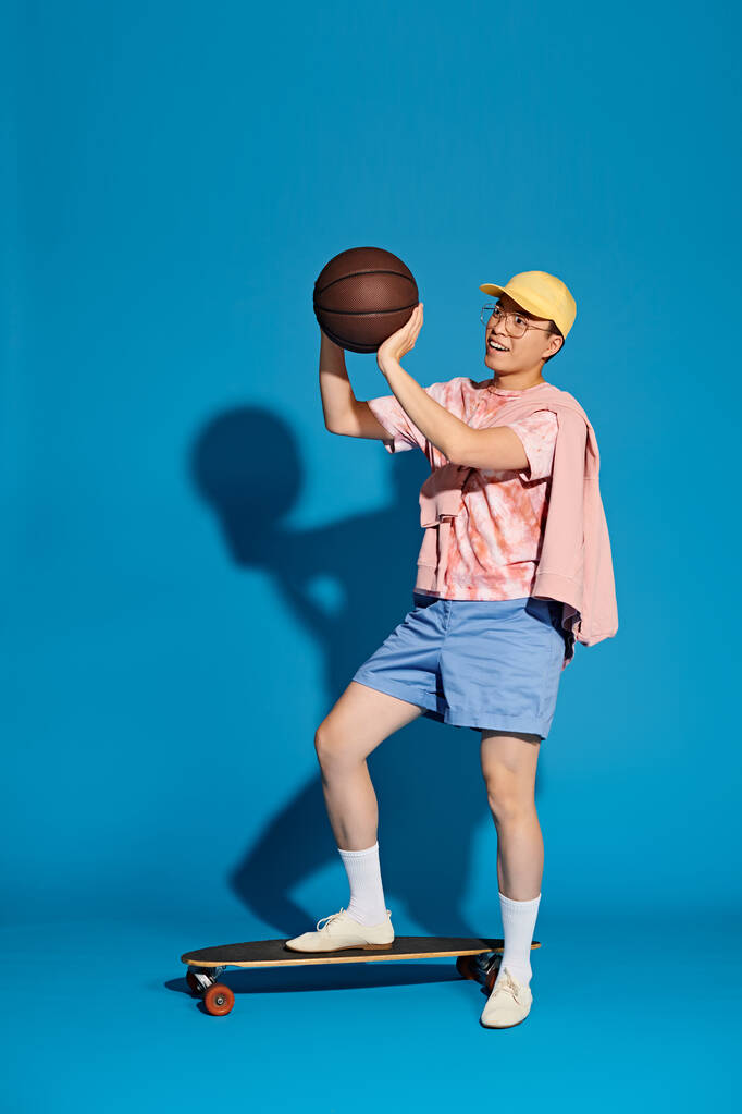 Stylowy mężczyzna trzymający koszykówkę podczas balansowania na deskorolce na niebieskim tle. - Zdjęcie, obraz
