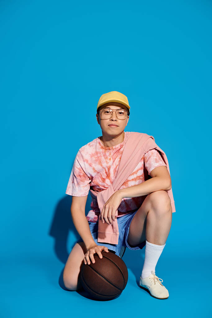 地面に座っているトレンディな服装のスタイリッシュな若い男,自信を持って青い背景にバスケットボールを保持. - 写真・画像